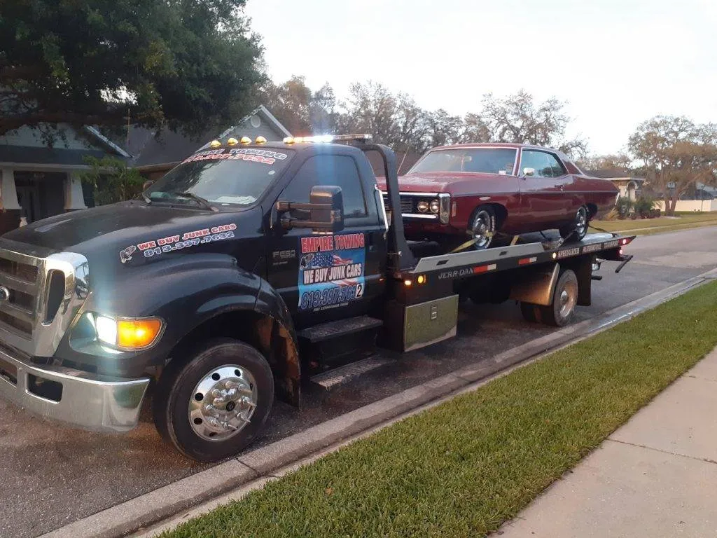 Headlights on, the Empire Towing flatbed carries a maroon classic car past a lawn